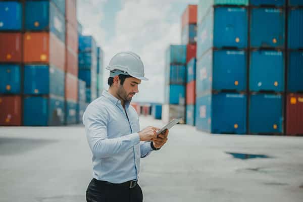 Engineer worker in casual suit standing in shipping container yard holding laptop with smile. Import and export product. Manufacturing transportation and global business concept.