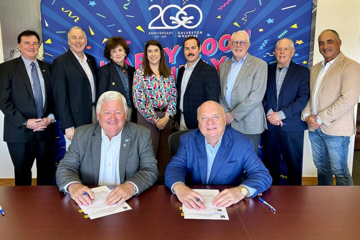 Group photo from the signing ceremony establishing a Galveston Wharves scholarship endowment for Texas A&M Galveston maritime students. Seated at a table are Port Director and CEO Rodger Rees and Board Chairman Vic Pierson signing documents. Standing behind them are representatives from the Texas A&M Foundation and members of the Galveston Wharves Board of Trustees, including Anna Hyatt, Tony Brown, Jeff Patterson, Sheila Lidstone, Erik Stramblad, Mayor Craig Brown, Richard Moore, and Willy Gonzalez.